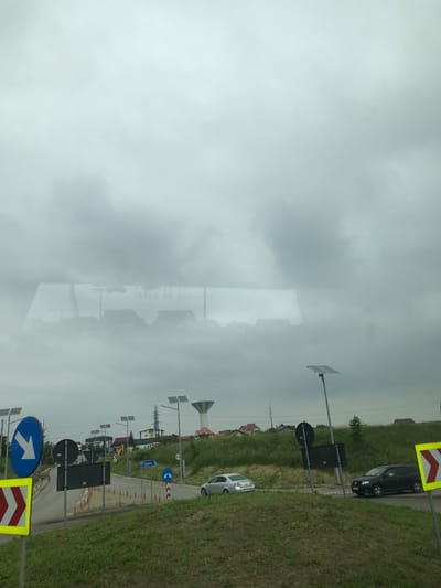 This image captures a street scene at a roundabout in Șcheia, Romania, on an overcast day. The perspective is from within a vehicle, evidenced by reflections in the upper portion of the frame.

The sky is uniformly gray, indicating dense cloud cover. In the foreground, a verdant grassy slope leads up to the road level. A yellow and red chevron sign, partially visible on the left, marks a curve or obstacle.

The midground is dominated by a paved roundabout with a central grassy island. A prominent blue circular sign with a white downward-pointing arrow is positioned on the left side of the roundabout. Adjacent to it are several black, rectangular signs, which are unreadable. Further along the roundabout, a blue rectangular sign clearly displays "209 Suceava 7" with a white right-pointing arrow, indicating direction and distance to Suceava. Another blue circular sign with two white curved arrows signifies the presence of a roundabout. Orange and white traffic cones delineate a section of the road. Multiple streetlights, each equipped with solar panels, are visible around the roundabout. Two vehicles are present: a silver sedan (possibly a Volkswagen Passat) is on the left side of the roundabout, and a dark SUV (possibly a Dacia Logan MCV) is on the right side.

The background features rolling hills covered in green vegetation, dotted with residential houses, many characterized by red roofs. A tall, white water tower with a conical roof stands out against the horizon on the right. Overhead utility lines are also visible. The upper area of the image contains a blurred, inverted reflection on glass, showing text that includes "ISSUE DE SECOURS" (French for "Emergency Exit") along with indistinct numbers and architectural outlines.