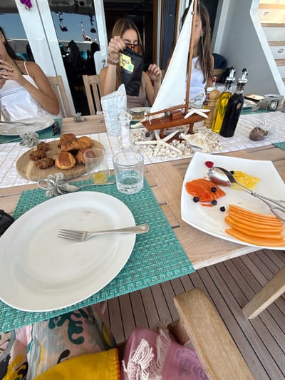 On a boat in Unknown, Mexico, a table is set with breakfast items. A woman in a white dress sits near a plate with pastries and a glass of juice. Another woman is holding a package of coffee or tea. On the table are bottles of oil and vinegar, a decorative sailboat, and a plate with fruit. A plate with a fork rests on a blue placemat. A wooden chair and a patterned pillow are also visible. The scene suggests a relaxed breakfast setting on a sunny day.