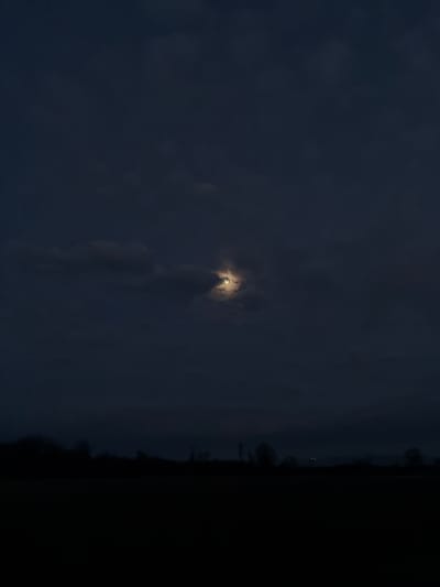 A blurry image reveals a small, bright moon centered in a dark, clouded sky. The lower half of the frame contains blurred cloud formations, while the right edge is a dense, nearly black mass, possibly trees or land against the night. Faint, distant lights are barely visible along the horizon.