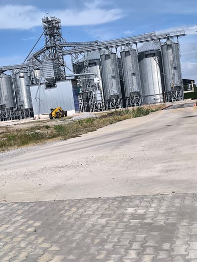 The image shows a large industrial facility, likely a grain silo complex, in Inovo, Bulgaria. The main structures are several tall, cylindrical metal silos arranged in a row, connected by a network of elevated conveyor belts and a central tower structure. The silos are made of corrugated metal and appear to be in good condition. A smaller, gray building is situated to the left of the silos, possibly an administrative office or processing plant. In the foreground, a yellow front-end loader is parked on a dirt and gravel area in front of the facility. The ground is a mix of dirt, gravel, and some patches of dry grass and small wildflowers. A paved area with interlocking concrete blocks is visible in the lower foreground. The sky is blue with scattered white clouds, suggesting fair weather. The overall scene depicts an active industrial site.