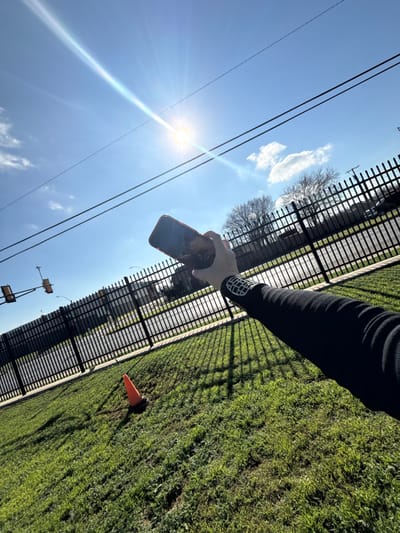 A sunny outdoor shot angled upwards toward the sky. A gloved hand holds a smartphone, its screen reflecting the surroundings. Power lines stretch across the bright blue sky, with a few wispy clouds visible. A black metal fence runs alongside a grassy area, where an orange traffic cone sits. The perspective is unusual, with the camera tilted, giving a distorted view of the landscape.
