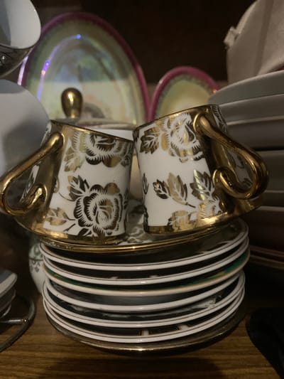 The image shows a close-up view of a stack of vintage fine china plates and two teacups with gold floral accents. The teacups are white with gold rose and leaf designs and have gold handles. They are placed on saucers which rest on top of a stack of plates. The plates are white with gold and black decorative rims, and some appear to have different colored bands. In the background, other pieces of china are visible, including a iridescent platter with a pink rim and a gold finial. The scene is set on a wooden surface, likely a shelf. The lighting is dim, creating shadows. The overall impression is of an antique collection displayed indoors. The location is not discernible from the visual information provided in the image.