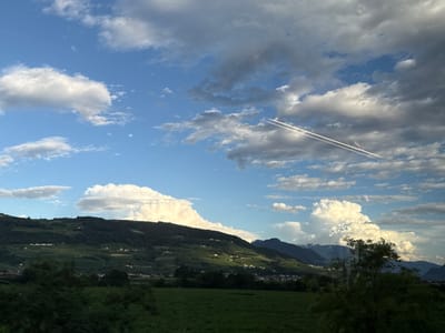 The image depicts a scenic view of rolling hills covered in vineyards under a partly cloudy sky. The sun is casting a warm glow on the landscape, highlighting the green terraces of the vineyards and scattered white houses. In the foreground, there are dark green trees and a lush green field. In the sky, two parallel contrails from an airplane are visible, streaking across the blue expanse. The location is Nave San Rocco, Italy, a region known for its agricultural beauty.