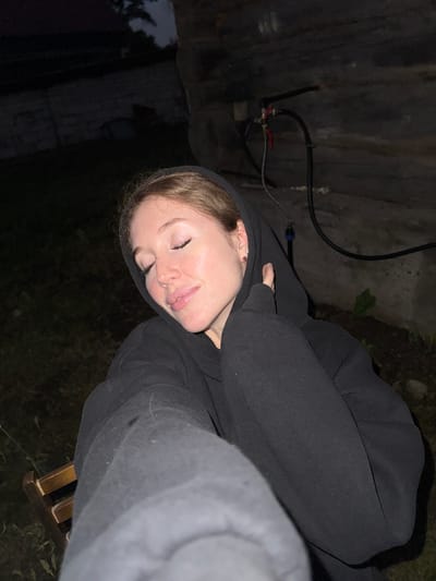 A young woman with auburn hair pulled back and wearing a black hooded sweatshirt is captured in a close-up, waist-up, low-angle selfie. Her eyes are closed, and her head is tilted back slightly, with her hands tucked into the hood of her sweatshirt, suggesting a relaxed or perhaps sleepy pose. The background is dark and somewhat out of focus, hinting at an outdoor setting at dusk or night. Visible in the background is a rough, stone-like wall with some pipes and hoses attached, and a patch of dark earth with some sparse vegetation. There is also a hint of a wooden chair on the left side of the frame. The overall lighting is dim, with the woman's face illuminated by a soft light source, possibly a flash or ambient light. The location is identified as Maksimovo, Russia.
