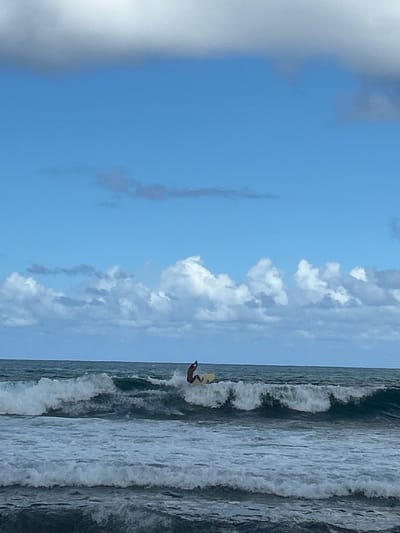 This image depicts an outdoor maritime scene in Le Moule, France. A single individual, dressed in dark attire, is positioned on a light-colored surfboard, actively riding a breaking wave in the midground. The person is crouched with arms bent and raised, appearing to maintain balance. The ocean features multiple waves, with white foam visible from breaking crests. The horizon line separates the dark blue-grey ocean from a light blue sky, which contains scattered white cumulus clouds and some darker grey cloud formations.