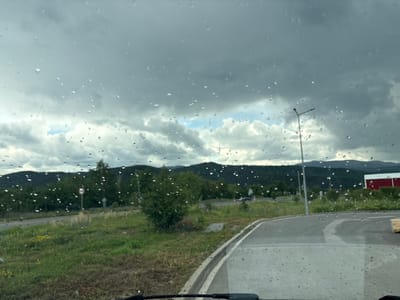 The image presents a view through a rain-covered vehicle windshield, indicating recent or ongoing precipitation. Numerous raindrops adhere to the windshield glass, causing minor visual distortion. The vehicle's dashboard or lower interior is visible at the bottom of the frame.

The environment is a semi-rural or industrial area under a sky dominated by dark grey clouds with intermittent patches of lighter grey and blue visible. A wet, paved road curves to the right in the midground. Adjacent to the left of the road is a grassy median containing green and dry vegetation, interspersed with yellow and purple wildflowers, bushes, and trees. A red and white circular traffic sign is positioned on the left, partially obscured by greenery. A grey street lamp pole with a cantilevered arm is situated on the right side of the road.

Dark green, forested hills delineate the horizon in the background. A red building with a white roof is partially visible through trees on the far right. No individuals are present. The location is Pirdop, Bulgaria.