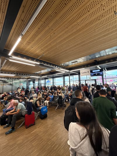The image depicts a crowded airport departure gate area, likely at Memmingen Airport in or near Künersberg, Germany, given the text on the blue bag. A large number of people are waiting, many seated on benches and others standing. The environment appears to be an indoor terminal with a wooden paneled ceiling featuring numerous perforations and modern fluorescent lighting. Large windows offer a view of an outdoor tarmac, where an aircraft with "WIZZ" branding is visible. A sign indicates "Gate 4". Among the people are individuals of various ages, dressed in casual travel attire. Luggage, including suitcases and backpacks, is present around the waiting passengers. The overall scene conveys a sense of anticipation and waiting, typical of an airport environment before boarding.