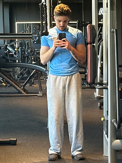 A single male individual is depicted standing full-length in what appears to be a gym in Jestetten, Germany. He has light brown hair, styled with volume, and a light beard. He wears a light blue short-sleeved t-shirt with lighter accents on the shoulders, loose-fitting light gray sweatpants, and dark athletic shoes. He is holding a black smartphone with multiple rear cameras in both hands at chest height, pointed towards himself, indicating a mirror selfie. A tattoo with text is visible on his right forearm. The environment consists of dark gray rubber flooring and various gym equipment in the background, including a multi-station cable machine with a visible weight stack and reddish-brown padded cushions on the right, and other metallic structures and workout apparatus on the left. A faint reflection of the letter "A" is visible on a mirrored surface in the upper right background.