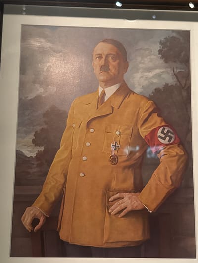 The media file is a photograph of a painting. The painting depicts a man in a tan uniform with a white shirt and tie. He has a mustache and is looking towards the viewer. On his left arm is an armband with a swastika symbol. He is also wearing medals on his uniform. The background of the painting is a misty, cloudy landscape, with trees visible. The man's right hand rests on his hip, while his left arm is bent and his hand is resting on his chest. The lighting in the photograph of the painting suggests an indoor setting, possibly a museum or gallery. There is no visible text in the image. The style of the painting appears to be realistic. The setting cues in the painting are ambiguous, but the swastika armband clearly indicates a connection to Nazi Germany. The emotion conveyed by the subject is stoic and unreadable.