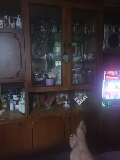 The image is an indoor shot of a wooden cabinet with glass doors, likely in a home. The cabinet contains various items, including what appears to be glassware, decorative objects, and toiletries on the shelves. To the right of the cabinet, a television screen displays a digital clock showing "12:17" and some brightly colored content. A speaker is positioned above the television. In the foreground, a person's bare feet are visible, suggesting they are reclining. One foot has dark red nail polish. A decorative item with the Russian words "Я ТЕБЯ" (I love you) and a heart is placed on a shelf below the cabinet. The overall impression is a casual, domestic setting. Without further context, the specific location in Pereleshinskoye, Russia, cannot be determined from the image alone, as it depicts a generic interior scene.