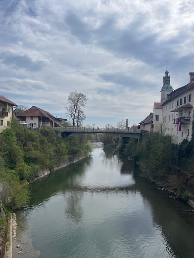 The image depicts the Poljanska Sora river flowing through Skofja Loka, Slovenia. The waterway is flanked by buildings and vegetation on both sides. A bridge spans the river in the distance. A prominent building with a tower is visible on the left bank, while buildings with red-tiled roofs are on the right. The sky is overcast with clouds.