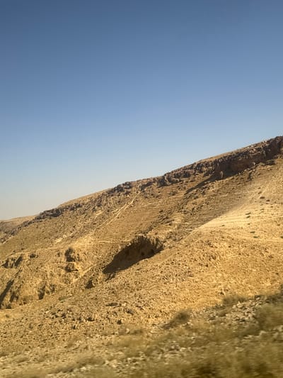 The image shows a rocky, arid cliff face under a clear blue sky. The cliff is a dominant feature, occupying the right two-thirds of the frame, and its textured surface displays various shades of brown and tan. There is a noticeable indentation or small cave on the cliff face, casting a shadow. The sky is a gradient of light blue, becoming lighter towards the horizon, suggesting it is daytime. The sun appears to be high, as evidenced by the shadows cast. The landscape appears barren and dry, typical of a desert or semi-arid region. There are no people or man-made objects visible in the image. The overall impression is one of natural, rugged terrain. The location is identified as Arad, Israel, which aligns with the arid environment depicted. The image is oriented vertically, with the cliff face running diagonally from the top right to the bottom left.