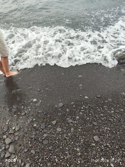 The image shows a close-up view of a pebble beach in Camogli, Italy, with the Ligurian Sea in the background. The tide is coming in, with a wave of white foam washing over the dark, wet pebbles and the feet of a person standing at the water's edge. The person is wearing light-colored shorts. The date and time stamp "31.07.2026 18:08" and "POCO F5" are visible in the lower corners of the image. The scene depicts a moment of interaction between a person and the sea on a rocky coastline.