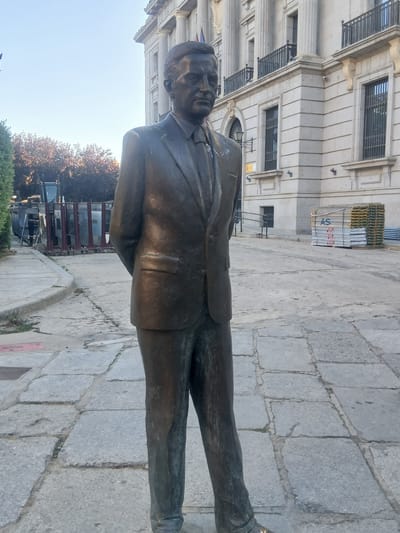 A full-body bronze statue of a man in a suit stands on a paved walkway in front of a stone building. The man is facing to the right of the frame with his hands clasped behind his back. The building behind him has a classical facade with columns, balconies, and barred windows. In the middle ground to the right of the building, a stack of scaffolding is visible. To the left of the statue, a sidewalk and a patch of grass are present, with a large construction dumpster and some portable barriers further back. The ground is paved with large, irregular stone slabs. The sky is clear and blue. This statue is located in the Plaza de Santa Ana in the city of Ávila, Spain.