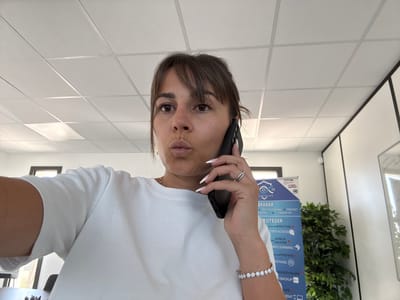 The image depicts a woman on the phone in an office environment. She is wearing a white t-shirt and appears to be making a "kissing face" gesture. A banner with text in French, possibly related to IT services, is visible in the background, along with a whiteboard. A potted plant is also present. The ceiling appears to be a drop ceiling with fluorescent lighting. The overall scene suggests an indoor office setting. The location is specified as Les Arcs, France.