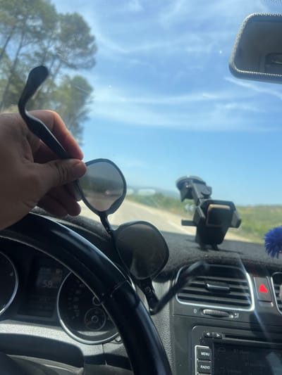 A hand holds sunglasses in a vehicle. The car's interior is visible, including the dashboard, steering wheel, and rearview mirror. Outside the car is sky and greenery. Derdara, Morocco cannot be confirmed based on visual evidence.