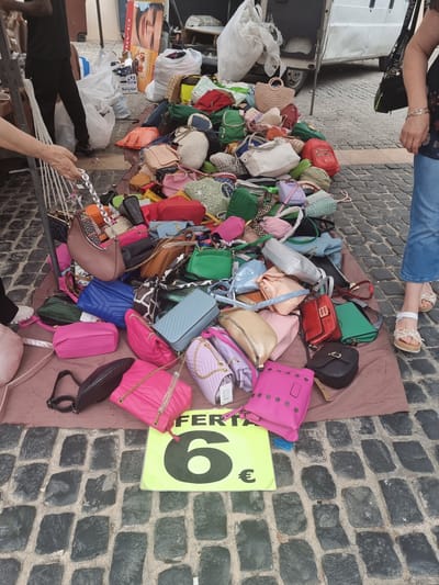 The image captures an outdoor market stall in Xàtiva, Spain, during daylight hours. A large collection of women's handbags and purses of various sizes, styles, and colors—including pink, green, red, blue, black, gold, and purple—is piled on a brown fabric spread on a cobbled street. Many bags feature chain straps, while others have fabric or faux leather straps.

Several individuals are present. A person's legs and feet, wearing jeans and sandals, are visible at the top, potentially browsing. Another person's hands are shown at the bottom left, holding a string of pearls or beads, possibly arranging merchandise. A third person's lower body is seen on the bottom right, near the bags.

Adjacent to the vibrant display of bags are white plastic bags, possibly containing other goods or packaging, and part of a white market van. A prominent yellow-green sign with bold black text reads "FERTA" (likely a shortened form of "Oferta," meaning 'sale' or 'offer') and a large "6€", indicating a price of 6 euros for items, presumably the handbags. A "CLARINS" product box is also visible amidst other items. The cobbled pavement confirms an outdoor market setting, suggesting a lively shopping experience.