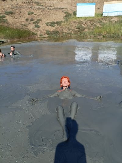 A group of people are relaxing in a mud bath in an outdoor setting. The water is a thick, grey mud, and the people are submerged to their chests or necks. In the foreground, a woman with short, red hair is lying back with her eyes closed, her arms outstretched. Her body is covered in mud, and she appears to be enjoying the experience. In the background, other people are also partially submerged in the mud, including a man holding a child.
The scene appears to be set during the daytime, with bright sunlight casting shadows. The surrounding landscape is dry and hilly, with sparse vegetation. Two informational signs are visible in the background, one on the left and one on the right, mounted on poles. The signs are white with blue and black text. The location is identified as Za Rodinu, Russia. The overall atmosphere is one of relaxation and well-being.