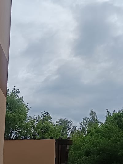 This is a close-up, vertical, eye-level shot of a wall and trees under a cloudy sky. The image appears to have been taken in Milovice, Czechia. The left side of the frame is dominated by the side of a building with a beige and brown facade. To the right of the building's facade, a low, tan-colored wall topped with black spikes is visible. Behind the wall, lush green trees fill the lower portion of the frame, extending upwards. The sky above is overcast with gray clouds, obscuring any hint of sunlight. No people are visible in the scene.