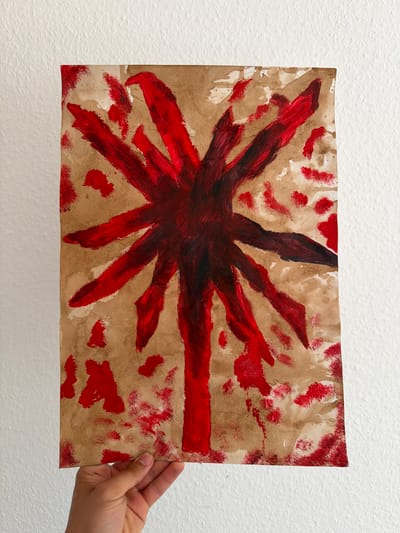 A close-up shot shows a child's hand holding a piece of art against a textured white wall. The artwork is on a light brown paper background, appearing somewhat creased and stained. The dominant element of the painting is a dark red, starburst-like shape in the center, with long, spiky strokes radiating outwards. There are also smaller, scattered red splotches and streaks across the paper, giving it an abstract and somewhat messy appearance. The child's hand, visible on the right side of the image, is gently holding the edge of the paper, suggesting the artwork is being presented or displayed. The lighting is even, and there are no obvious external environmental cues like weather or time of day. The location context of Nienburg/Weser, Germany, is noted but not visually represented in the image itself. There is no visible text on the artwork or in the scene.