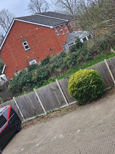 The scene depicts a residential area during daylight hours under an overcast sky, located within the Cherwell District, United Kingdom. The specific city cannot be determined from the visual information provided. No individuals are present in the image.
In the foreground, a grey and tan herringbone-patterned paved surface extends across the lower portion of the frame. A black van with a distinct red horizontal stripe along its side and black multi-spoke wheels is partially visible, parked on the paved surface towards the left. To the right, a manicured, spherical-shaped green shrub grows on sparse ground cover, adjacent to a weathered wooden panel fence.
This fence, characterized by vertical wooden panels and light-colored concrete posts, runs horizontally across the midground. Immediately behind the fence lies a dense and unkempt strip of green and brown shrubbery and overgrown vegetation.
In the background, a two-story residential building with red brick exterior walls and a dark grey tiled pitched roof is prominently featured. The building includes multiple white-framed windows; specifically, a vertical rectangular window on the upper floor and a smaller horizontal window on the lower floor are visible on one side, while three smaller vertical windows are grouped together on the upper floor of a recessed section. Another residential structure with a lighter grey pitched roof is partially visible further in the background to the right. Several bare deciduous trees with intricate branch patterns are silhouetted against the overcast sky at the top of the frame.