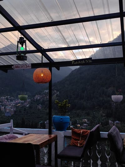 The image depicts a rooftop seating area in Old Manali, with a view of a hillside village and a dense forest. In the foreground, a wooden table is set with chairs. To the left, a white bench is adorned with a pink cushion. To the right, two chairs are covered with dark fabric, and an orange cushion with gold accents is placed on a seat. A blue pot containing a plant sits on a ledge.
Above, a transparent roof with metal framing covers the area. A vintage-style lantern with a green bulb hangs from the frame, and a woven spherical lamp with a lit bulb illuminates the space. Two signs are visible: one points to "LAHAUL SPITI 50 KM," and another reads "Rishikesh 513 KM." Hanging planters, one green and one white with a pink flower, are suspended from chains.
The background reveals a multi-layered view of buildings nestled into a steep, tree-covered mountain slope, with lights visible in some structures, suggesting the setting is likely dusk or early evening. The sky above the mountains is cloudy, with hints of sunset colors.