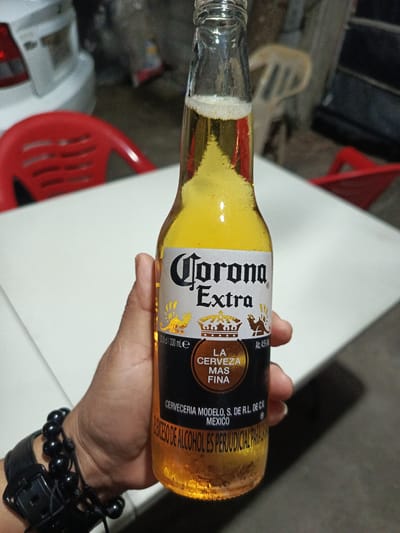 A hand holds a bottle of Corona Extra beer. The bottle is yellow-amber, with a white label bearing the beer's name and a golden crown. The label also has text in Spanish, "LA CERVEZA MAS FINA." The bottle has condensation. The hand wearing a black beaded bracelet and watch. The background includes a white table, red chairs, and the rear of a white car. The location, Quevedo, Ecuador is not explicitly identifiable in the image.