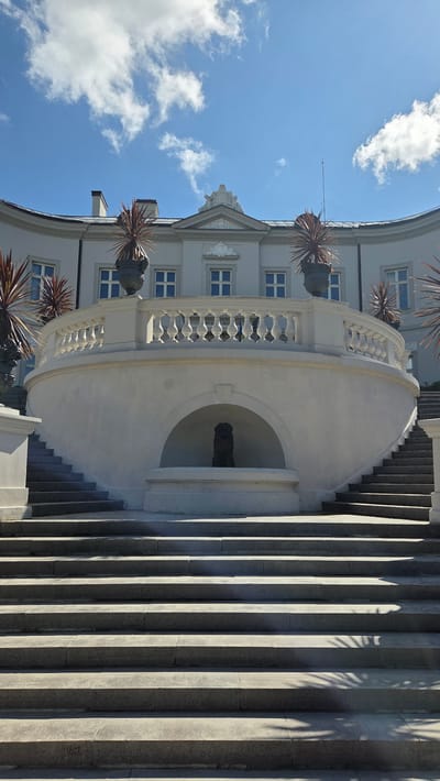 A low-angle, vertically oriented shot captures an ornate white building with a curved facade, set against a bright blue sky with scattered white clouds. The building features multiple windows and decorative architectural elements. In the foreground, a grand, curved staircase ascends along the side of the building, with a white balustrade and planters containing spiky, reddish-brown foliage. At the base of the staircase, within a semicircular alcove, a dark sculpture is visible. The steps of the staircase are made of stone, casting long shadows from an unseen light source, suggesting it is daytime. The image conveys a sense of grandeur and classical architecture. This scene is located in Palanga, Lithuania.