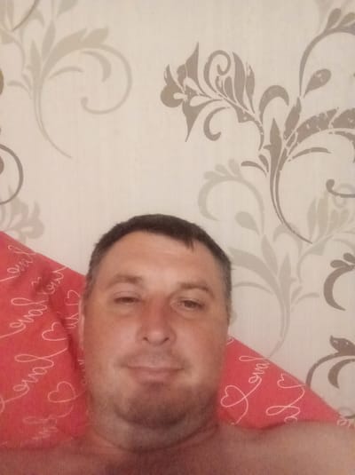 A middle-aged man with short, dark hair and a light complexion is shown from the chest up, looking directly at the camera. He has a short beard and is wearing a red pillow with white script and heart designs. The pillow is positioned under his head and to his left. The background consists of patterned wallpaper with light beige and grey floral designs. The image appears to be a selfie taken indoors.