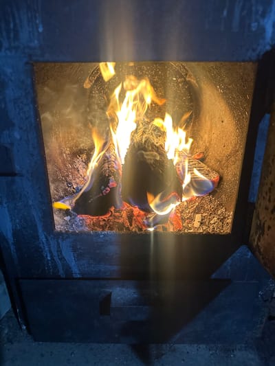 The image shows a close-up view of a wood-burning stove with burning logs inside. The flames are a vibrant mix of orange, yellow, and blue, casting a warm glow on the surrounding ash and soot-covered interior. Smoke is visibly rising from the fire. The exterior of the stove appears to be made of dark, heavy metal, with some textured surfaces and what looks like a lower compartment or door. The overall environment is dark, suggesting an indoor setting, possibly a home or rustic cabin, in the city of Karachino, Russia.