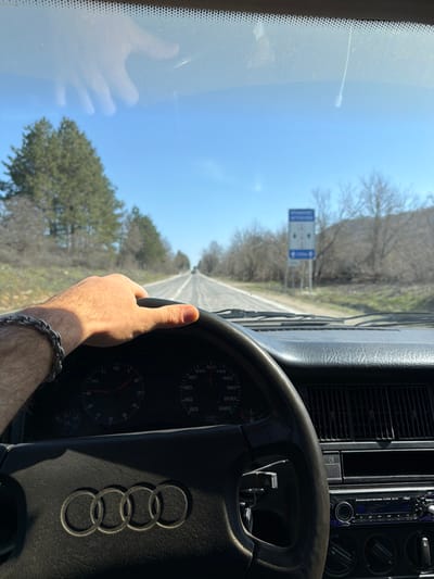 The image presents a first-person view from the interior of a vehicle traveling on a two-lane asphalt road under a clear, bright blue sky. A light-skinned male's right hand, adorned with a silver-colored braided bracelet on the wrist, grips the black Audi steering wheel at approximately the 10 o'clock position. The Audi logo, consisting of four interconnected rings, is prominent on the steering wheel's center.

Behind the steering wheel, the vehicle's instrument cluster is visible, showing a tachometer with a needle near zero and a speedometer indicating approximately 60 km/h. The dark dashboard features visible air vents and a partially obscured aftermarket stereo unit with blue illumination. A faint reflection of a hand is visible on the upper left portion of the windshield.

The road ahead is straight and appears somewhat worn, with white dashed lane markings. Sparse vegetation and dirt shoulders border the road. On the left side, a cluster of evergreen trees is prominent, while the right side features deciduous trees with bare branches, suggesting late winter or early spring. Further along the right side of the road, a rectangular blue road sign is visible. The sign displays white text in Cyrillic, reading "ВНИМАНИЕ!" (ATTENTION!) at the top, followed by "МОСТОВЕ" (BRIDGES) and "ЯМИ" (POTHOLES), indicating a warning for road conditions. Distant hills are visible on the horizon, particularly to the right. The scene depicts travel on a rural road in the vicinity of Malinovo, Bulgaria, as specified.