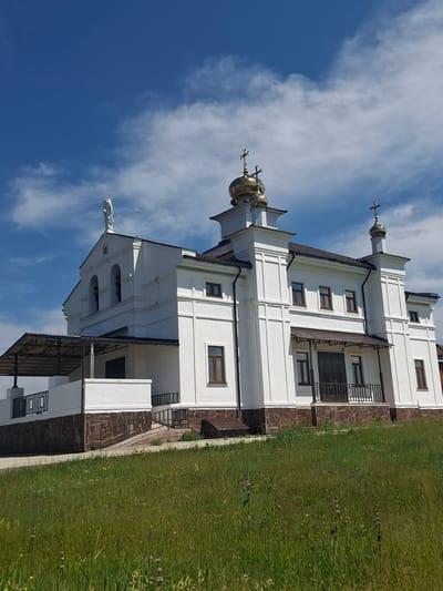 A low-angle, medium shot captures a white church with golden domes against a bright blue sky with scattered white clouds. The church features a statue of a figure standing atop its roof and a bell tower with arched openings. A ramp leads to an entrance porch with a dark door. The base of the church is clad in dark brown stone. The foreground is a grassy slope with wildflowers. The scene is static and depicts a building in a rural or semi-rural setting. The location is identified as the city of Bayanday, Russia.