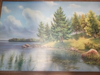 This is a landscape painting of a tranquil lake with a wooded area on the opposite bank. The foreground depicts the calm surface of the lake, with reeds and lily pads growing in the shallow water. Large, rounded rocks are scattered along the shore and in the water, adding texture and visual interest.
The middle ground features a gentle slope leading up to a stand of trees. Prominent evergreen trees dominate the center of the composition, with deciduous trees showing lighter green and yellow foliage towards the right. The trees are rendered with detail, suggesting a lush and healthy environment. Sunlight appears to be filtering through the trees, creating dappled light and shadow on the grassy embankment.
In the distance, the opposite bank of the lake is lined with a dense arrangement of trees, creating a soft, hazy horizon. The sky is a pale blue with large, fluffy white clouds, suggesting a pleasant day. The overall mood of the painting is serene and peaceful, evoking a sense of natural beauty and quietude. There is no visible text in the image. The time of day appears to be late morning or early afternoon, with bright but diffused light. The location is likely a natural setting with a lake and surrounding woodland.