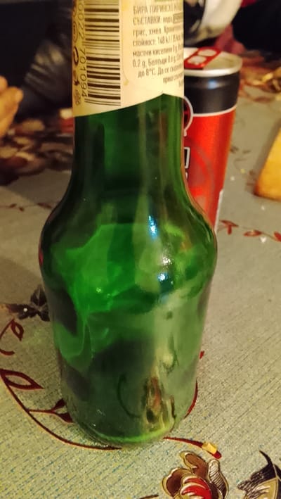 The scene depicts a tabletop arrangement in an indoor setting within Neunkirchen, Germany. In the foreground, a translucent green glass bottle, partially filled with a dark liquid, stands upright. A light-colored paper label encircles the bottle's neck, displaying a visible barcode (0022 701094) and text written in Cyrillic script, including "БИРА ПИРИНСКО" (Beer Pirinsko) and nutritional information. To the right and slightly behind the bottle, a red and black aluminum beverage can with a silver top is visible. Both containers rest on a light-colored tablecloth patterned with floral motifs in red, brown, and yellow hues. Blurred objects are present in the distant background; no individuals are depicted.