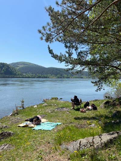 This image captures a tranquil outdoor scene by a wide river in Sliznevo, Russia, under a clear, bright blue sky. The perspective is from a slightly elevated position on the riverbank, looking across the water towards a distant, forested landscape.
In the foreground, a verdant grassy bank slopes gently towards the river, dotted with rocks and vibrant yellow wildflowers. Two people are present on this bank, enjoying the sunny weather. One individual, dressed in dark clothing and a cap, is seated on a small green folding chair near the water's edge, appearing to be focused on an object held in their hands, possibly a book or a mobile device. A dark backpack and other bags rest on the grass beside them. Further along the bank to the right, a second person is visible, lying or reclining on a light-colored blanket, partially covered by a blue and green patterned blanket or towel. They are dressed in a light-colored top and dark trousers.
The wide river flows calmly across the middle ground, its surface reflecting the daylight. On the far bank, densely forested mountains or hills rise, stretching into the distance under the clear sky. Branches of a large pine tree, with its characteristic needles, frame the upper left corner of the image, contributing to the natural ambiance. The strong, direct sunlight suggests it is mid-day. The overall scene conveys a sense of peaceful leisure and connection with nature. No visible text is present in the image.