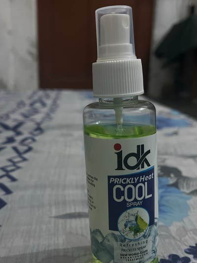 A close-up, vertical shot shows a bottle of "IDK Prickly Heat Cool Spray" with green liquid inside, resting on a patterned surface. The bottle has a white spray nozzle and cap. The label features the brand name "idk," the text "PRICKLY Heat," "COOL SPRAY," and an illustration of lime and mint leaves with ice. Below this, smaller text reads "Refreshing PRICKLES SPRAY NEW WORLD-CLASS BREAKTHROUGH TREATMENT." The background is softly blurred, showing a brown wall or furniture and a patterned tablecloth or surface with blue and white designs. No people are visible. The image appears to be taken indoors, likely in a home environment in Kabal, Pakistan.
