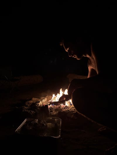 At night in the Arava Tichona Regional Council, Israel, an individual is observed crouched or seated on dark, uneven ground, tending to a campfire. The person, viewed in profile from the right, is shirtless with facial hair, illuminated primarily by the orange and blue flames of the fire. Their right hand holds a stick, extending it into the burning embers. To their immediate left, a rectangular aluminum foil pan rests on the ground, containing several foil-wrapped bundles, likely food items. Wisps of smoke rise from the fire. In the distant left background, a dark, partially visible folding chair is present. The surrounding environment is predominantly dark, indicating an outdoor, unlit area.