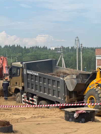 In Chulman, Russia, a construction site is depicted. A large gold SHACMAN dump truck is positioned, its bed filled with dirt. An orange excavator is parked to the left of the truck, partially visible. A worker in a blue uniform and orange helmet stands near the excavator, facing the truck. Red and white striped caution tape is stretched across the foreground, indicating a restricted area. A yellow wheel loader is situated to the right of the dump truck, with its bucket extended over the truck's bed, appearing to be in the process of dumping dirt. In the background, a dense line of green trees stretches across the horizon, with a few buildings and power lines visible above the treeline. The ground is sandy and uneven, typical of a construction environment.