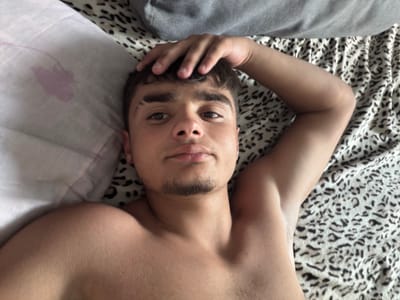 A young man is lying in bed, looking at the camera. He has dark hair and is wearing a grey t-shirt. The bed has a leopard-print comforter and a pink pillow. The room appears to be a bedroom. There is no information about the location in the city of Avdarma, Moldova.