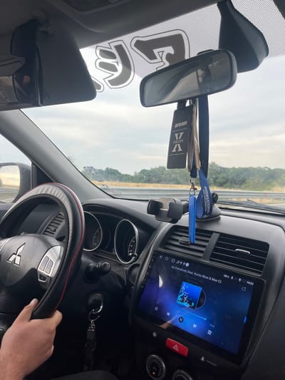 The image shows the interior of a car, likely a Mitsubishi, being driven on a road. The driver's hand is visible on the steering wheel. The dashboard features a multimedia system displaying song information and playback controls, an air vent, and a car air freshener hanging from the rearview mirror. The windshield shows a view of a rural landscape with trees and a cloudy sky. The scene is captured from the passenger's perspective, looking forward. There is no indication of Mirovo, Bulgaria in the image.