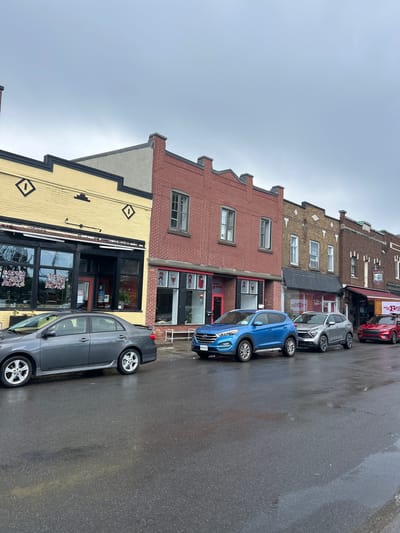 The image depicts a street scene in Sainte-Anne-de-Bellevue, Canada. The street is wet, indicating recent rain or damp conditions. Several buildings with storefronts line the street, featuring a variety of architectural styles and brickwork.
In the foreground, a gray sedan is parked, followed by a blue SUV, a silver SUV, and a red SUV. The blue SUV has a license plate visible that says "MONTREAL". The red SUV has a sign on its side that partially reads "WORLD CUP". Storefront signs are visible on the buildings, including one with "cafe" visible on the yellow building. Windows on the storefronts reveal the interiors of the businesses, some of which appear to be shops or cafes. The sky is overcast and gray. No people are visible in the image.