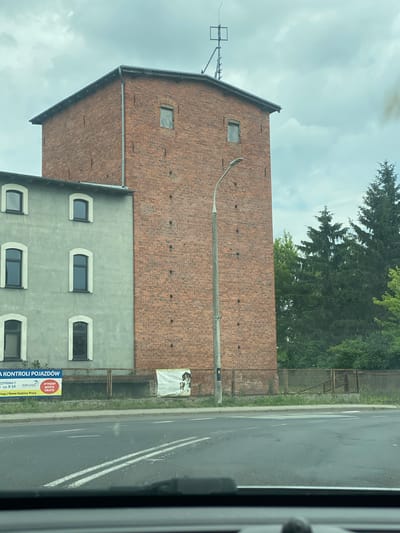 The image captures an exterior view of a brick building in Miłosław, Poland. The building features a prominent, tall brick tower with several small, rectangular windows. Adjacent to the tower is a lighter-colored building with multiple arched windows. In front of the buildings, a street is visible with double white lines painted on the asphalt. Behind the buildings, a chain-link fence separates the property from an area with grass and trees, including several tall evergreen trees. A lamppost stands in front of the brick tower. Two signs are visible attached to the fence: one with text in Polish and a graphic that appears to be related to vehicle inspection, and another larger banner with an image of people. The sky above is overcast.