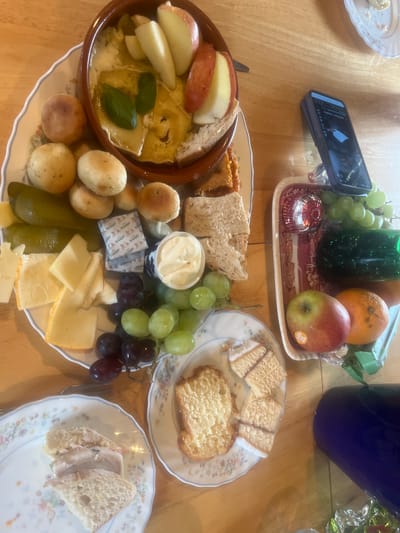 The image shows a wooden table laden with a variety of food items, suggesting a social gathering or meal. A smartphone rests on a decorative tray containing grapes, an orange, and an apple. Several plates display bread, cakes, cheese, and what appears to be a baked cheese dish garnished with apple slices and basil. The items are arranged on floral-patterned plates. The environment is an indoor setting with natural light. The city of Golspie, United Kingdom, cannot be confirmed from this image alone.