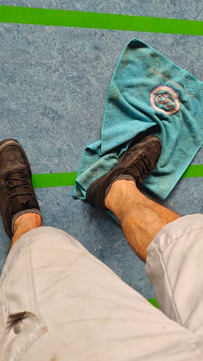 This media file shows a top-down view of a person's legs and feet on a blue, speckled floor with a thin green line marking a boundary. The person is wearing black athletic shoes and light-colored pants, which appear to be work pants given some dirt stains. A folded turquoise microfiber towel with a circular embroidered logo is placed on the floor next to one of the shoes. The lighting suggests it is daytime. The logo on the towel contains red and white lettering that is difficult to fully decipher but includes "CC" prominently. The scene appears to be indoors, possibly in a gymnasium or similar facility, given the floor surface and the green line, which could be a court marking. No specific activity is depicted, but the presence of work pants and a towel might suggest a break during physical labor or sports. The location context provided is Maintal, Germany.