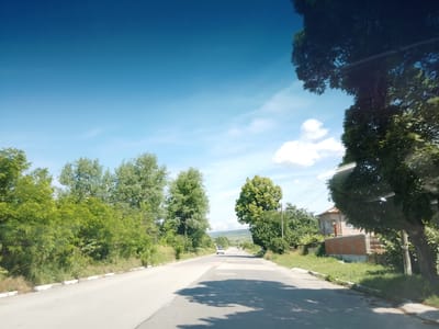The image depicts a road in Mirovo, Bulgaria, on a clear, sunny day. The road appears to be a two-lane asphalt road with a white line marking its edge on the left side. Trees and vegetation line both sides of the road, with some taller trees casting shadows on the asphalt. In the distance, a white car is visible traveling in the same direction as the camera. Further down the road, another vehicle can be seen. To the right, a house with a red-tiled roof and a brick wall is partially visible behind a dense growth of trees. The sky is a vibrant blue with scattered white clouds, and a power line is visible running through the scene. The overall impression is one of a rural or suburban setting on a pleasant day.