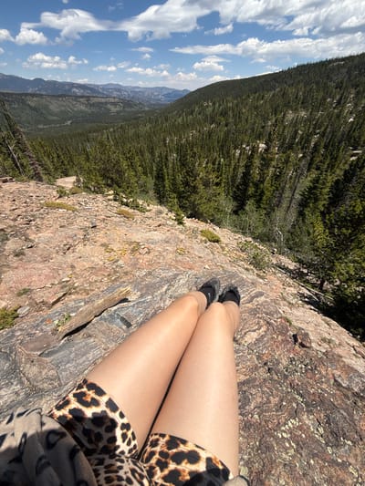 This image depicts a person's legs and feet resting on a rocky outcrop overlooking a vast, forested valley. The person is wearing leopard-print shorts and dark athletic shoes. The valley below is densely covered with evergreen trees, and in the distance, a range of mountains stretches across the horizon under a partly cloudy blue sky. The rocky surface in the foreground is rough and appears to be a natural geological formation. The scene suggests an outdoor recreational activity, likely hiking or enjoying a scenic view. The specific location in Estes Park, United States, is not explicitly identifiable from the visual information alone.
