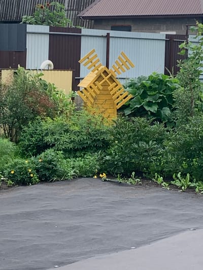 The image depicts a decorative wooden windmill, painted yellow, positioned in a garden area. The windmill is surrounded by lush green bushes and some yellow flowers. In the background, a corrugated metal fence, a brick wall, and parts of a building with a brown roof are visible. A section of dark gray ground cover is present in the foreground. The scene appears to be a residential or garden setting, likely in Dubnik, Russia, given the architectural styles and vegetation. There are no people or animals present in the image, and no actions or interactions are taking place.