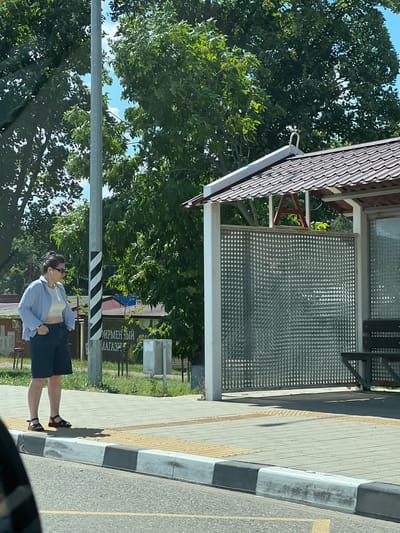 The image captures a street scene in Belozyorny, Russia, featuring a woman standing on a sidewalk next to a bus stop. The woman, wearing a light blue unbuttoned shirt, a white crop top, and dark shorts, has her hands in her pockets. She is also wearing sunglasses and has dark hair pulled back. The bus stop has a brown tiled roof, white pillars, and a perforated metal wall. A wooden bench is visible under the bus stop shelter. The sidewalk is paved with light gray square tiles and bordered by a tactile paving system. A striped pole with black and white bands stands to the left of the woman. In the background, trees with lush green foliage are visible, along with buildings and a sign with Russian text. The overall environment suggests a sunny day in an urban or semi-urban setting.