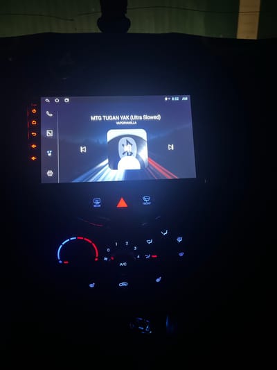 The image shows a car's dashboard at night. The infotainment system displays the song "MTG TUGAN YAK (Ultra Slowed)" by VAPORVANILLA. The climate control panel is illuminated, with settings visible for fan speed, temperature, and air conditioning. The picture appears to be taken in the dark. There is no indication of the location being in the city of Бизяки, Russia.