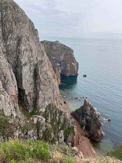 This image presents a top-down view of a rocky coastline, likely located in a coastal region. The scene is dominated by various shades of grey and brown rocks, some of which are partially covered with green vegetation. The water is calm and a muted grey-blue, with some areas appearing more transparent near the shoreline, revealing rocky formations below. There is a small beach with reddish-brown pebbles visible near the rocks. A rock formation is seen extending into the sea, featuring a small archway. The sky is overcast. The location is Russkiy Island, Russia.